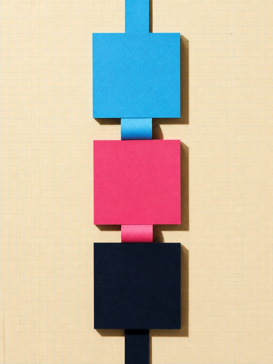 Three square paper panels — sky blue, rose pink and ink navy — stacked vertically and joined by folded paper strips, on cream graph paper, each casting a hard shadow. A strip enters above the top panel and leaves below the bottom one.